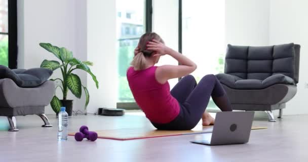 Fit женщина накачивает пресс, делая эффективные упражнения на коврик рядом с открытым ноутбуком в современных помещениях. Женщина в спортивной одежде практикует фитнес онлайн с тренером - Кадры, видео