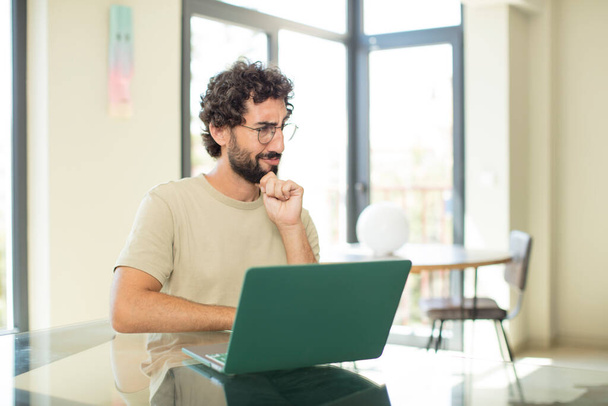 young adult bearded man with a laptop feeling confused or doubting, concentrating on an idea, thinking hard, looking to copy space on side - Foto, Imagem