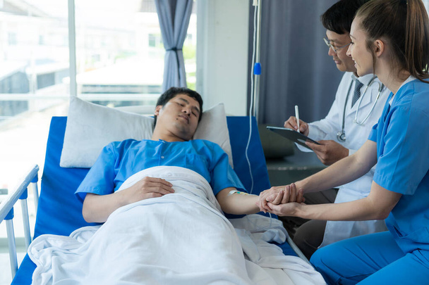 Δύο γιατροί που φροντίζουν έναν ασθενή ξαπλωμένο σε ένα κρεβάτι σε ένα δωμάτιο νοσοκομείου με δύο νοσοκόμες, έναν υπάλληλο υγειονομικής περίθαλψης που παρέχει υποστήριξη και συμβουλεύεται έναν ασθενή στο κρεβάτι - Φωτογραφία, εικόνα