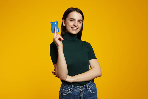 Young smiling happy fun caucasian woman wearing green shirt, hold in hand mock up of credit bank card isolated on plain yellow background - Photo, Image