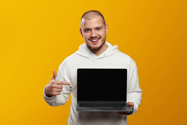 Giovane hipster barbuto sorridente con cappuccio bianco che punta il dito sul computer su sfondo giallo. - Foto, immagini