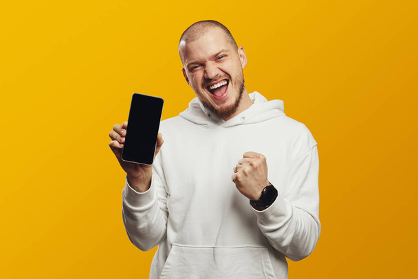 Excited young man screaming and showing empty screen smartphone while celebrating victory with raised fist, isolated over yellow background - Foto, Bild