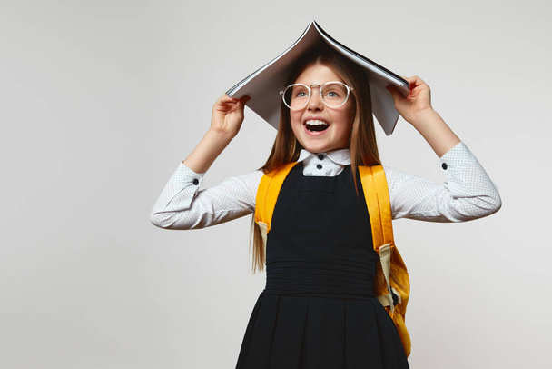 Surprised schoolgirl with eyeglasses wearing school uniform and yellow backpack yelling and covering head with book against white background - Photo, Image