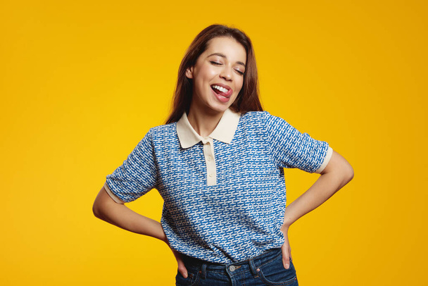 Portrait of beautiful woman holding hands on waist while sticking her tongue out isolated over yellow background - Valokuva, kuva