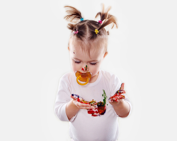 Het babymeisje met een fopspeen in gouache bevuild handen en shirt geïsoleerd - Foto, afbeelding