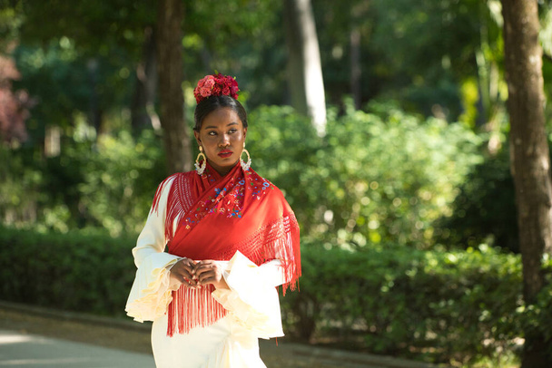 Une jeune femme noire déguisée en danseuse de flamenco se tient dans un célèbre parc à côté d'un étang de canards à Séville, en Espagne. Elle porte une robe beige avec des volants et châle rouge et des fleurs dans ses cheveux - Photo, image