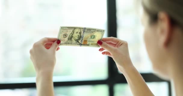 Woman with red manicure checks authenticity of dollar banknote holding bill against sunny light from window. Fake money concept. Criminal liability - Footage, Video