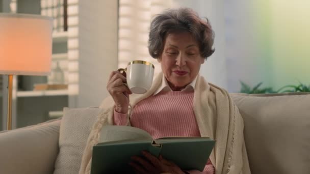 Szczęśliwy uśmiech zainspirowany białej starej kobiety pokryte kocem na kanapie picia kawy herbaty z filiżanki czytania książki uśmiech atmosferyczny spokój starsza pani starsza pani emeryt czytać napój w domu kanapa - Materiał filmowy, wideo