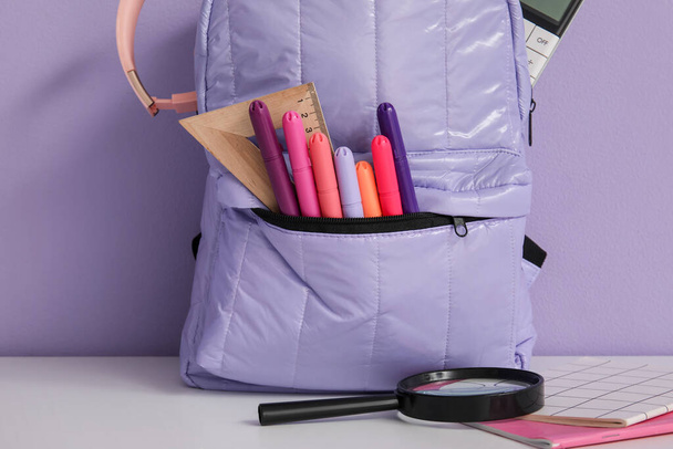Школьный рюкзак с различными канцелярскими принадлежностями и наушниками на белом столе возле фиолетовой стены - Фото, изображение