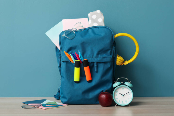 Bunter Schulrucksack mit verschiedenen Papeterie und Wecker auf Holztisch in der Nähe der blauen Wand - Foto, Bild