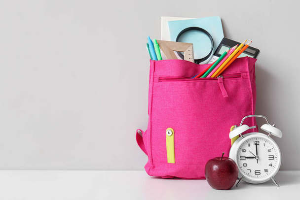 Zaino scuola rosa con diversi articoli di cancelleria, sveglia e mela fresca sul tavolo bianco vicino alla parete grigia - Foto, immagini