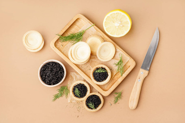 Wooden board of tasty tartlets with black caviar on beige background - Фото, изображение