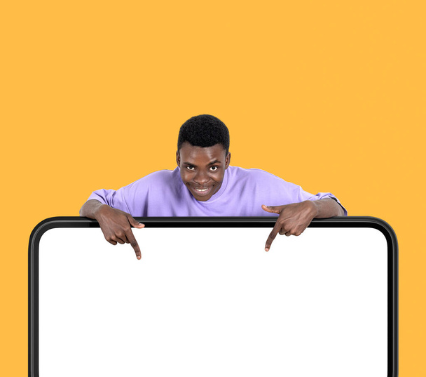 Black american man smiling, finger pointing down at big mock up blank digital screen on yellow background. Concept of subscribe and follow - Fotoğraf, Görsel