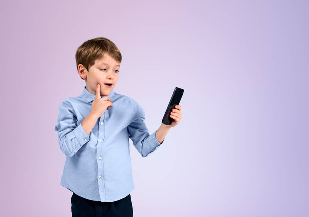 Child boy holding phone with ponder look, finger to cheek on empty copy space lilac background. Concept of social media and e-learning - Photo, Image