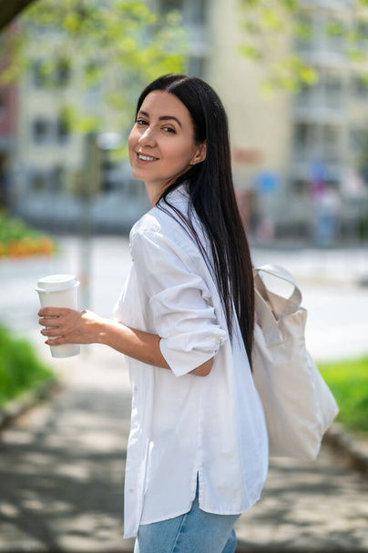Jovem feliz bonita com saco branco em pé na rua da cidade, beber café em papel, acordar na cidade. - Foto, Imagem