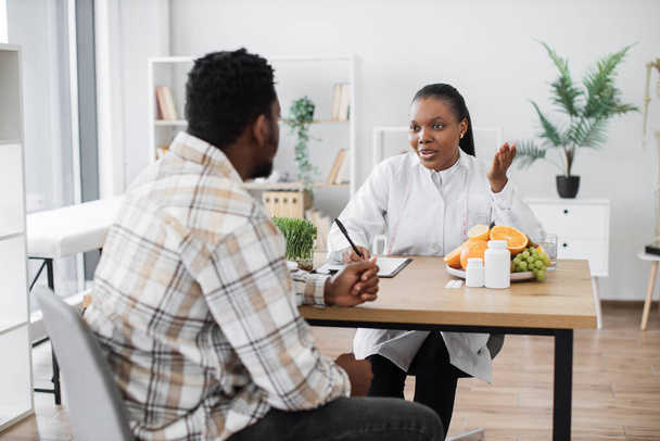 Focused multiethnic woman in doctors coat talking to middle-aged man in consulting room of hospital. Professional expert in nutrition providing personalized guidance to male client on food habits. - Photo, Image