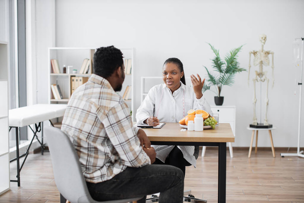 Focused multiethnic woman in doctors coat talking to middle-aged man in consulting room of hospital. Professional expert in nutrition providing personalized guidance to male client on food habits. - 写真・画像