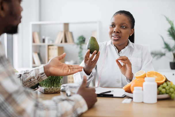 Beautiful multicultural woman with fresh avocado in hand chatting with bearded man in consulting room. Experienced medical specialist giving advice on improving diet by abundance of nutrients. - Fotoğraf, Görsel