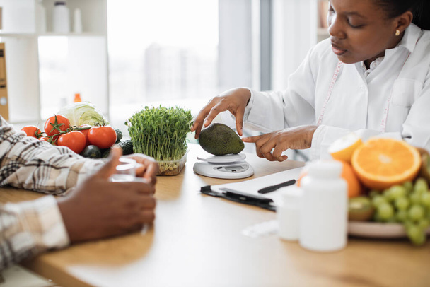 Beyaz önlüklü Afro-Amerikan diyetisyen hasta randevusu sırasında taze avokadoyu beslenme ölçeğine koyuyor. Kadın profesyonel akıllı teknolojilerle kalorilerin doğru ölçümlerini alıyor.. - Fotoğraf, Görsel