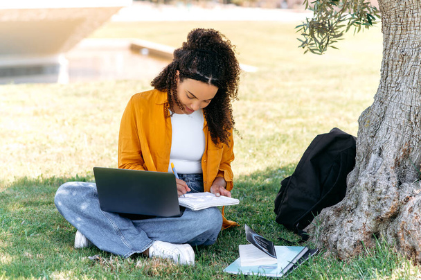 Learning concept. Modern stylish positive mixed race girl student, sitting on the grass near the tree, with a laptop and notepad, doing homework, listening to an online lecture, taking notes, smile - Photo, image