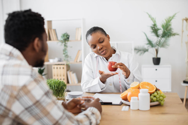 Female nutritionist holding apple with measuring tape while sitting at office desk in front of adult patient. Attractive multiracial woman in white coat promoting healthy eating with slimming foods. - Foto, Imagem