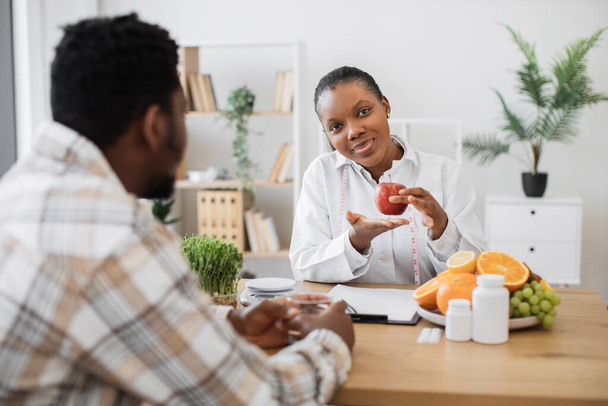 Female nutritionist holding apple with measuring tape while sitting at office desk in front of adult patient. Attractive multiracial woman in white coat promoting healthy eating with slimming foods. - Foto, Bild