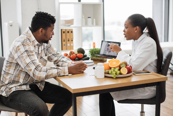 African american specialist in nutrition holding tablet with gastrointestinal tract on screen while counseling man at work. Focused woman explaining client about food intolerance during appointment. - 写真・画像