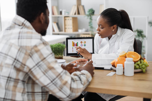 Over-shoulder view of african american woman analyzing patients nutrition data on digital tablet in consulting room. Efficient dietitian assessing health needs of male client in medical centre. - 写真・画像