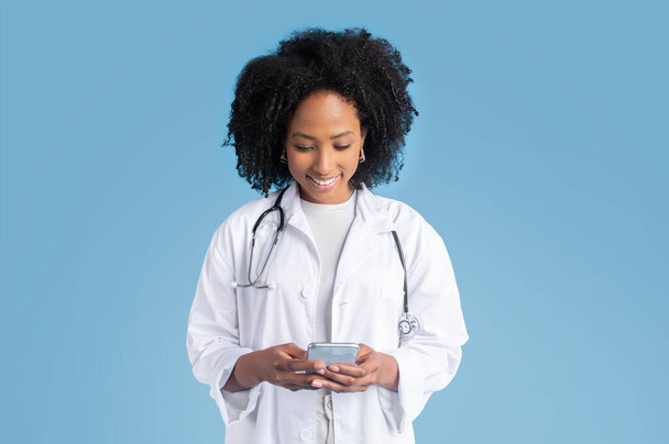 Heureuse jeune femme noire médecin thérapeute heureuse en manteau blanc tapant sur smartphone, bavardant, isolée sur fond de studio bleu. App pour la médecine, soins de santé avec gadget, message - Photo, image
