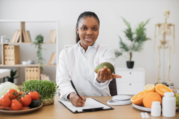 Portret van een lachende multiculturele dame in een witte jas met avocado in de hand poserend aan het bureau. Vrolijk expert in voedsel en voeding selecteren van voedsel voor speciale voeding in artsen werkplek. - Foto, afbeelding