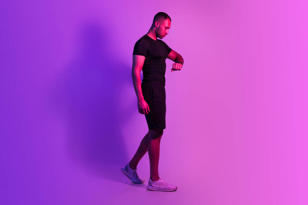 Workout Time. Sporty African American Man Walking Watching At Fitness Smartwatch On Wrist, Checking His Heartbeat And Burnt Calories After Sport Training Activities On Purple Studio Background - Foto, imagen