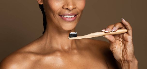 Dental health. Smiling black middle aged woman holding toothbrush with toothpaste, female ready to brush her teeth, standing over brown background, panorama, cropped, closeup - Photo, Image