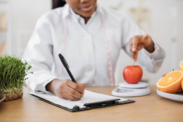 Close up view of health practitioner putting red fruit on digital device for weighing while recording information on paper. Multiethnic nutrition specialist achieving goals with smart technologies. - 写真・画像