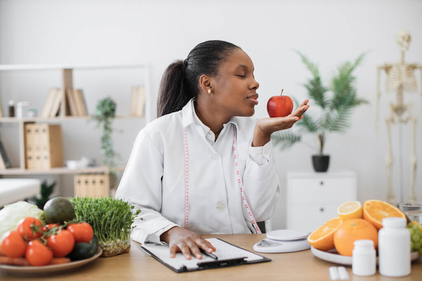 Charming adult lady with measuring tape smelling red apple on palm while noting down nutritional value on paper. Multicultural dietitian in white coat providing with cooking tips with fresh fruits. - 写真・画像