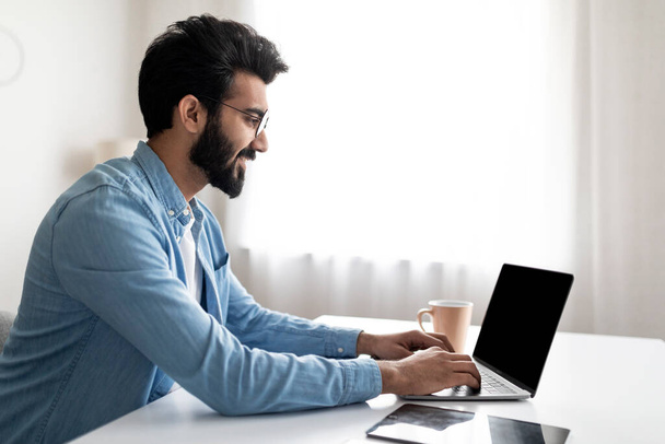 Homem indiano sorridente trabalhando no laptop com tela preta em branco no Home Office, jovem Freelancer masculino oriental sentado na mesa e digitando no computador, desfrutando de educação à distância e trabalho remoto - Foto, Imagem