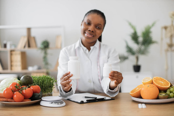 Focus on pills containers with vitamin supplements being held by multicultural woman at office desk. Medical specialist in white coat giving recommendations about filling nutrition gaps in diet. - Photo, image