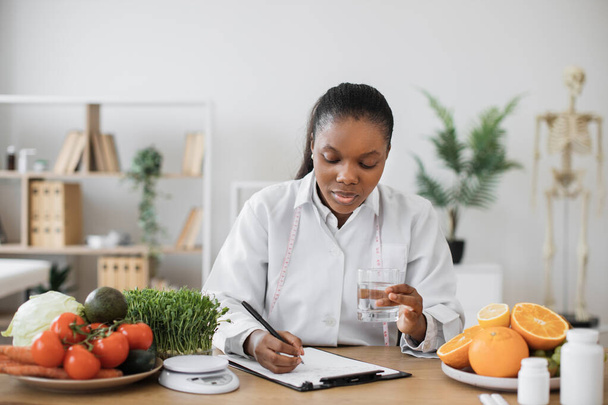 Thoughtful multiethnic lady with drink in hand making notes of diet changes while staying in doctors office. Experienced professional correcting amount of fluid for suppressing appetite with meals. - Foto, imagen