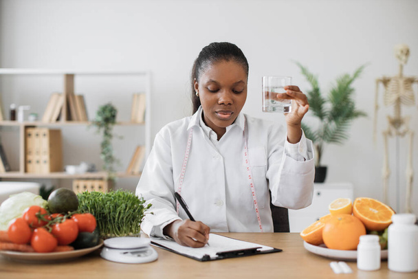 Thoughtful multiethnic lady with drink in hand making notes of diet changes while staying in doctors office. Experienced professional correcting amount of fluid for suppressing appetite with meals. - Photo, image