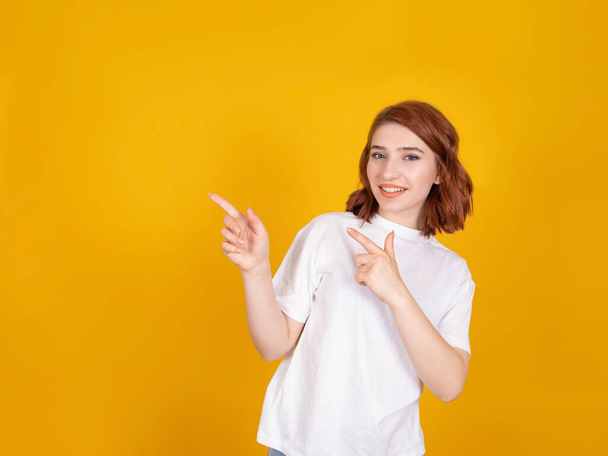 Pointing finger into blank space, smiling red hair caucasian girl wearing white shirt and pointing finger into blank space. Attractive beautiful lady standing over yellow background, copy space. - Foto, afbeelding