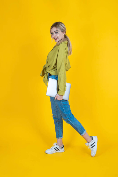 Holding closed laptop, full body side view young blonde woman holding closed laptop. Wearing jeans and green shirt, isolated yellow studio background. Walking, going, IT professional lifestyle concept - Photo, image