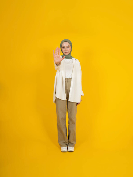 Muslim woman doing stop sign with palm of the hand. Freedom of religion, don't touch my head scarf. Warning expression negative, serious gesture. Full body front view girl standing yellow background. - Photo, image