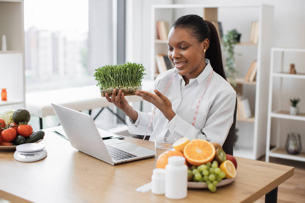 Smiling african american woman introducing microgreens during online conversation with client via laptop webcam. Modern nutrition professional advising young vegetable sprouts for healthy eating. - Foto, Bild