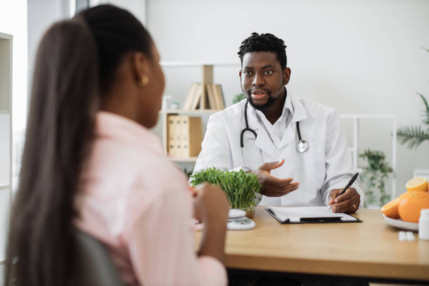 Confident man with stethoscope writing on paper clipboard during conversation with multiracial woman in office. Serious nutrition expert making notes of patients medical history for proper meal plan. - 写真・画像