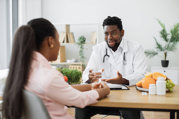 Attractive bearded man in doctors coat gesturing at glass with fluid while meeting with multiethnic patient. Smiling nutritionist recommending clean water as help in reducing calorie intake on diet. - 写真・画像