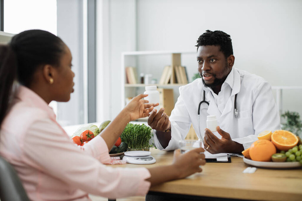 Handsome african american man in white coat giving pills bottle to charming woman in consulting room of hospital. Male nutrition professional improving overall health with dietary supplements. - Photo, image