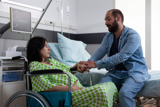 Kocası tekerlekli sandalyede oturan hamile kadını rahatlatıyor ve sezaryen ameliyatından önce ebeveynlik hakkında konuşuyor. Doğum kliniğinde bekleyen genç ebeveynler. Tıp konsepti - Fotoğraf, Görsel
