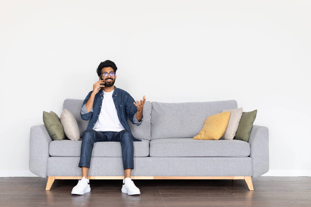 Concept de communication. Joyeux joyeux beau jeune homme arabe portant des lunettes assis sur un canapé confortable dans le salon, parlant au téléphone à la maison, regardant l'espace de copie, geste et sourire - Photo, image