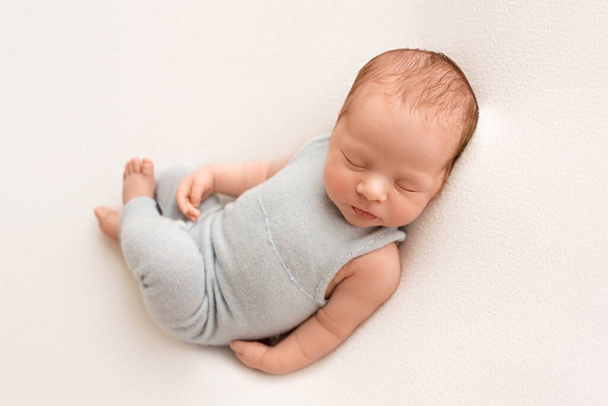 Un lindo niño recién nacido en los primeros días de vida duerme en un mono azul sobre un fondo de tela blanca. Foto de alta calidad - Foto, imagen