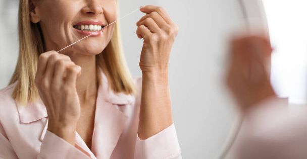Cropped Shot Of Smiling Middle Aged Woman Using Dental Floss At Home, Happy Mature Female Making Oral Care, Standing Near Mirror In Bathroom, Cleansing Teeth, Doing Daily Hygiene Routine, Closeup - Foto, Imagen