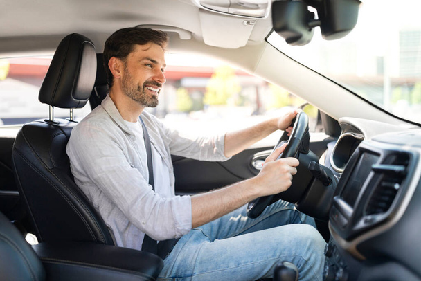 Happy cheerful handsome bearded young man wearing casual outfit driver sitting in front seat, holding auto steering wheel, driving his brand new nice car and smiling, copy space - Фото, изображение